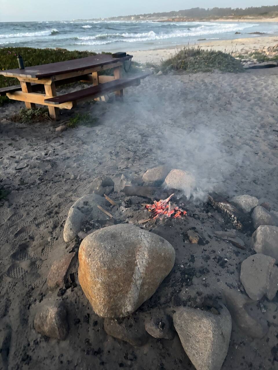 A campfire burning low in a stone-ringed pit on the beach at dusk, picnic table nearby, waves rolling in behind.