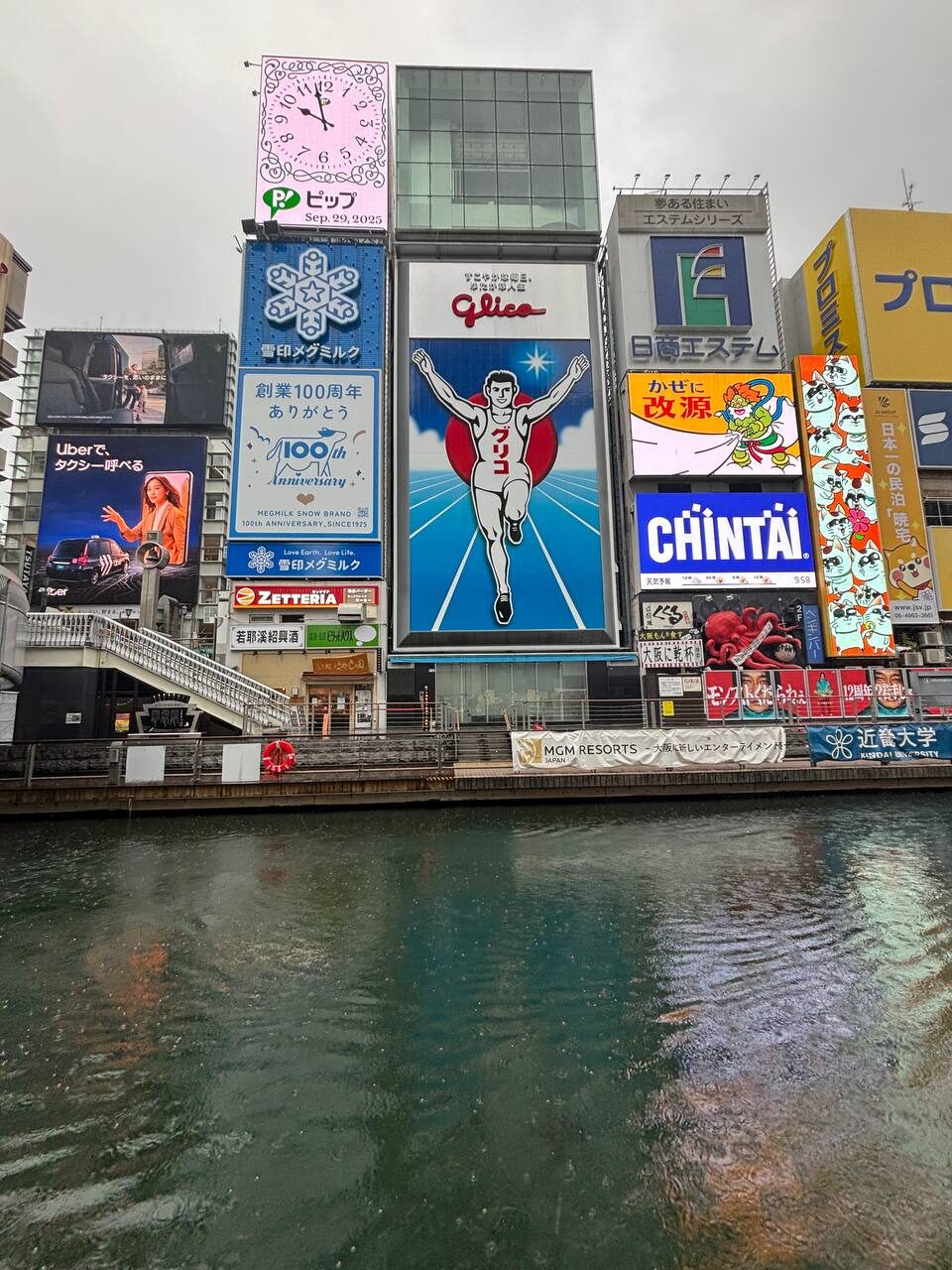 The Glico running man sign and surrounding billboards along the Dōtonbori canal in Osaka, reflected in the water on a rainy afternoon.